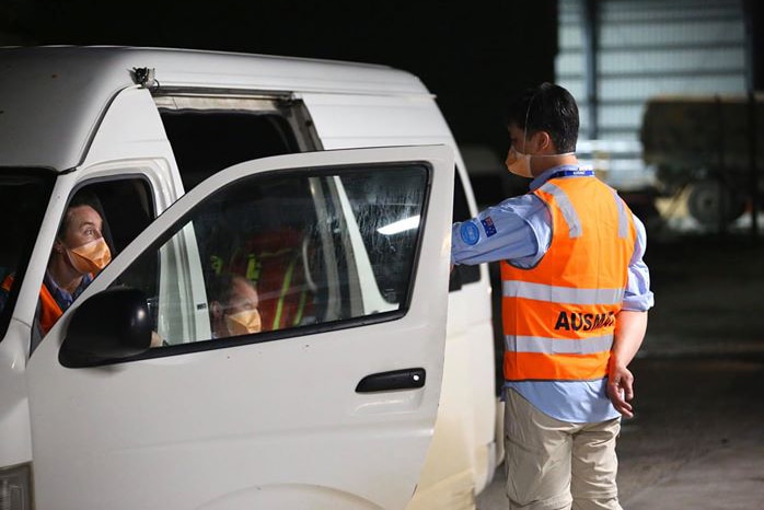 An AUSMAT medical worker stands next to a white van wearing a hi-vis vest with a colleague sitting in the car at night.
