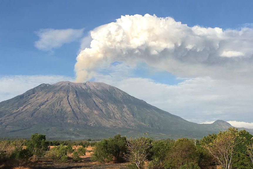 A volcano in the distance with a big white cloud of smoke coming out the top against a blue sky.