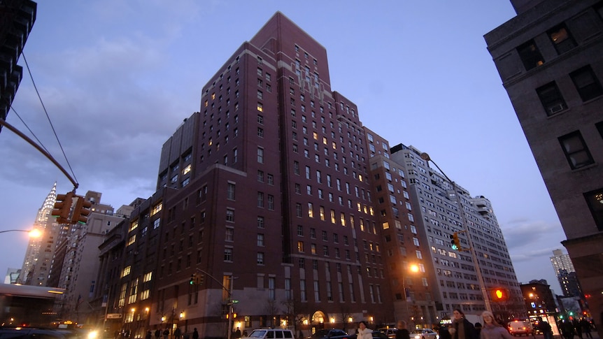 An imposing-looking reddish building, featureless except for glowing light inside some windows, on a New York block corner.