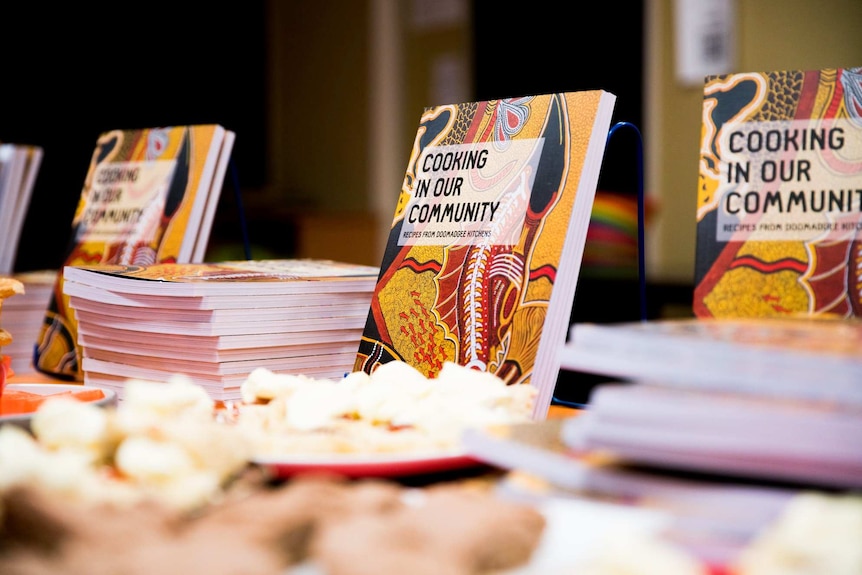 The Doomadgee cookbook, Cooking in Our Community: Recipes from Doomadgee Kitchens, is proudly propped up behind  plates of food.
