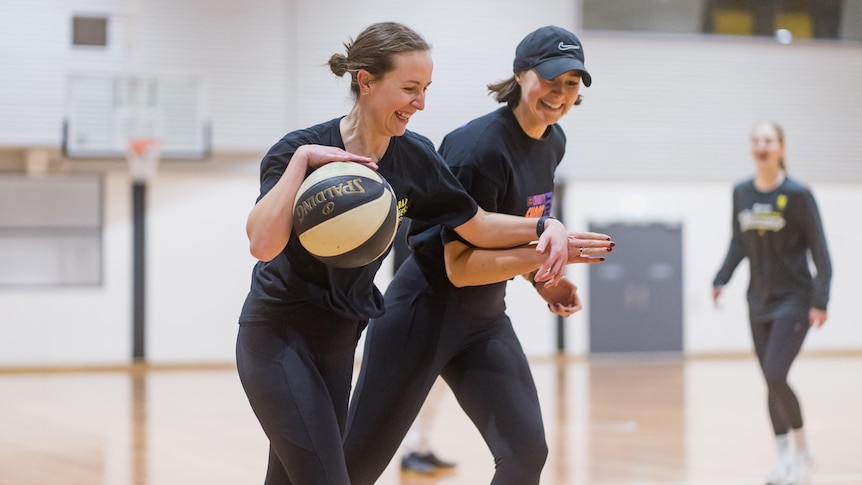 Two women on a basketball court. One is holding the ball and smiles playfully as she pushes a defender away.