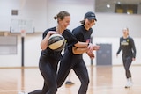 Two women on a basketball court. One is holding the ball and smiles playfully as she pushes a defender away.