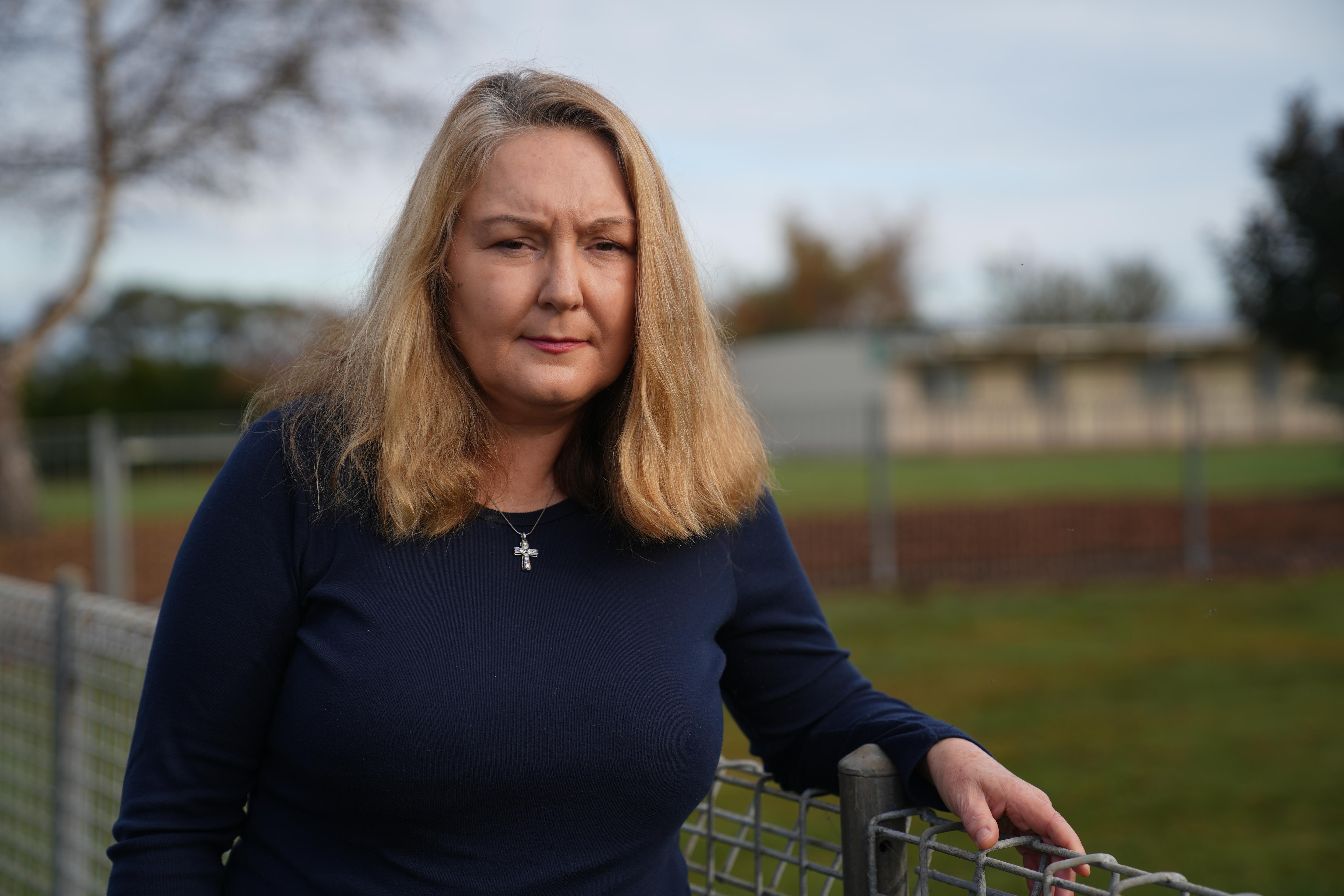She has straight blonde hair and wears a cross around her neck, as she stands by the school fence in morning light