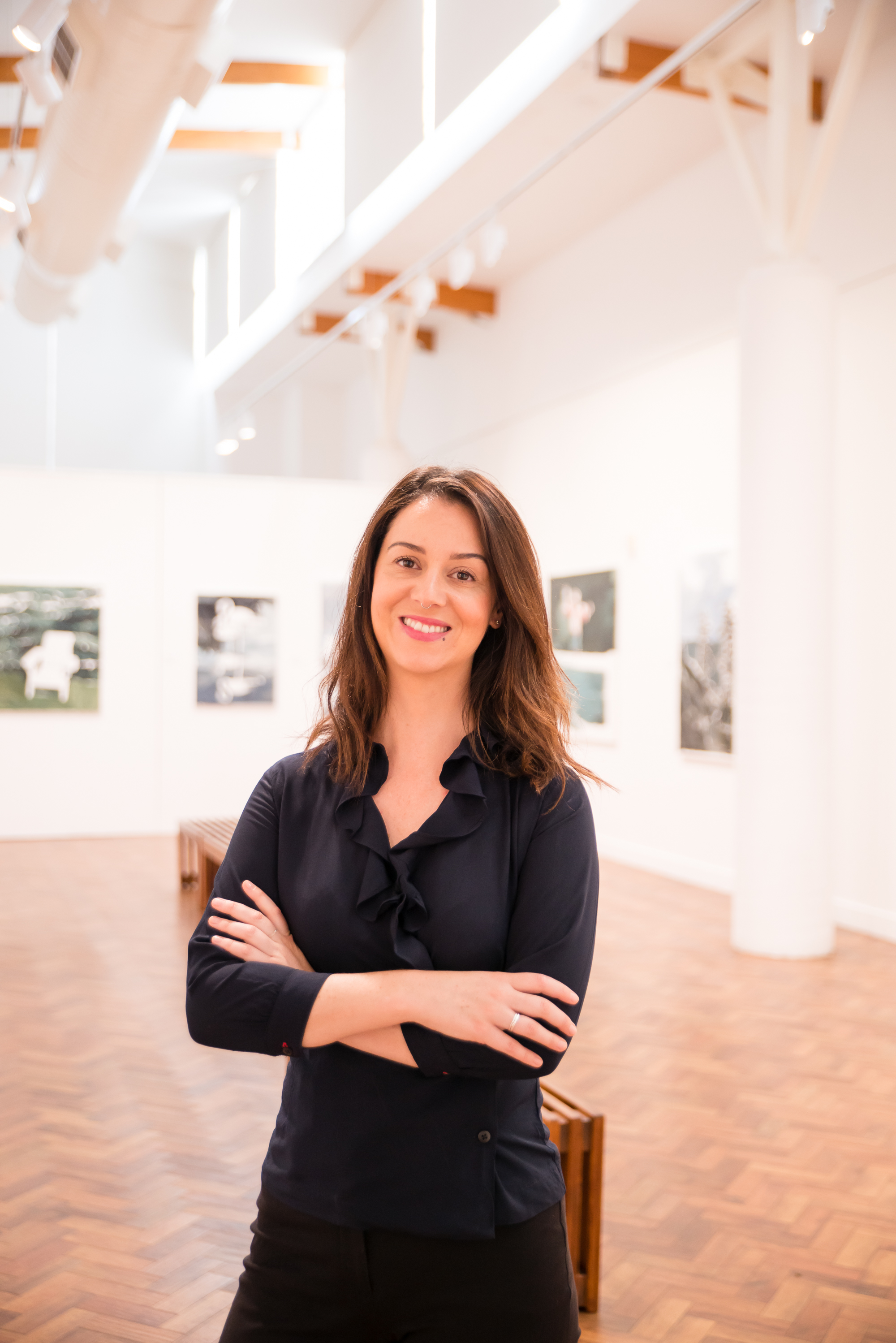 A 30-something woman with brown hair stands in a gallery space wearing all black. Paintings can be seen on the walls behind her.