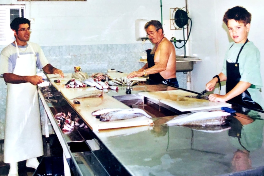 Older man in middle, middle aged man on left and young boy on right all with aprons filleting fish at a bench