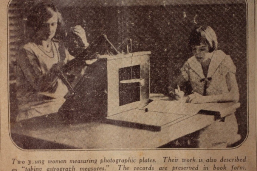 Newspaper clipping showing two women sitting at a desk analysing photographic glass plates and taking down notes in a logbook.