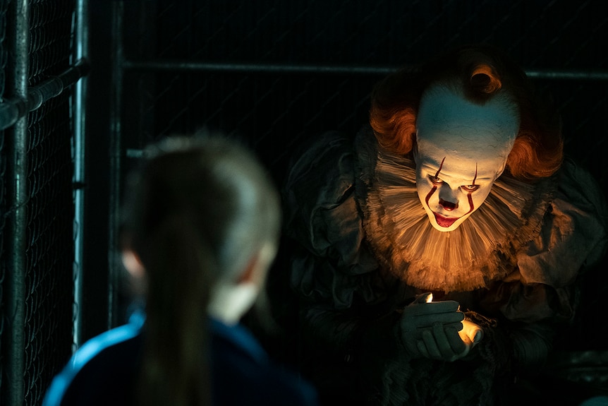 A small child looks at scary looking clown figure whose face is illuminated by a candle in their hands.