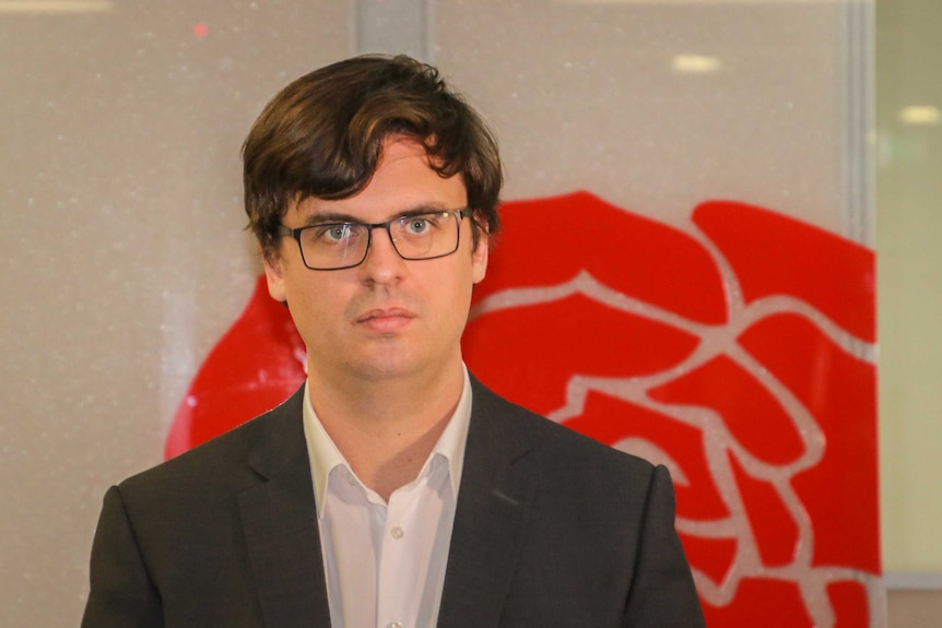 Assistant secretary of Unions WA Owen Whittle stands wearing a suit and glasses against a wall.