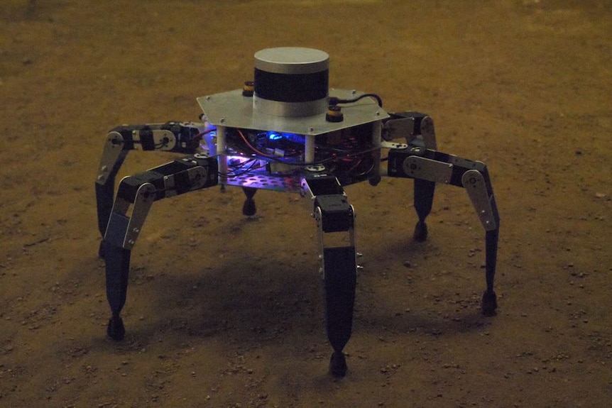 A robot shaped like a hexagon with spider-like legs stands in a dimly lit cave. Its body has purple flashing lights in it