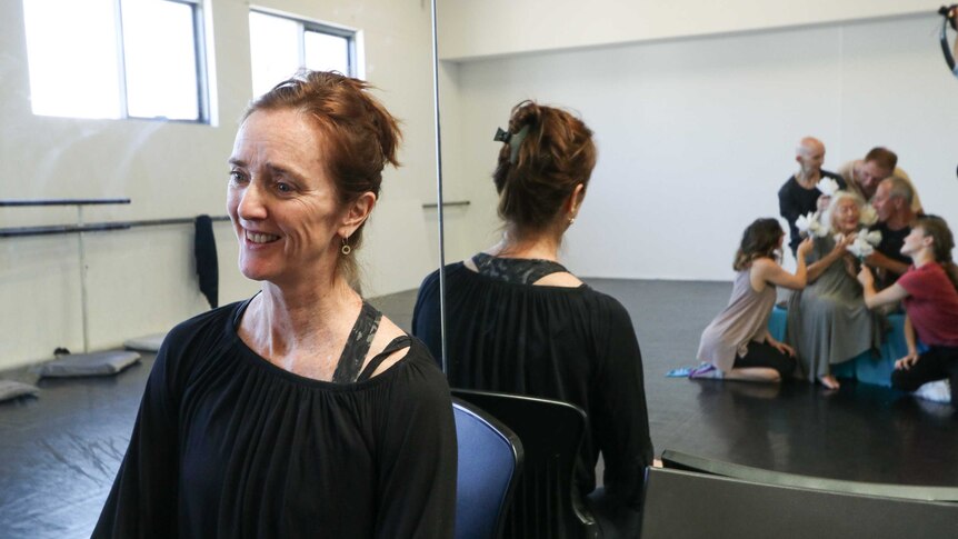 Sue Healey sitting with her back to a mirror reflecting an image of Eileen Kramer surrounded by dancers in rehearsal