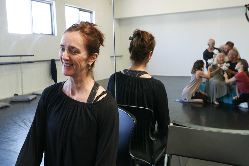 Sue Healey sitting with her back to a mirror reflecting an image of Eileen Kramer surrounded by dancers in rehearsal