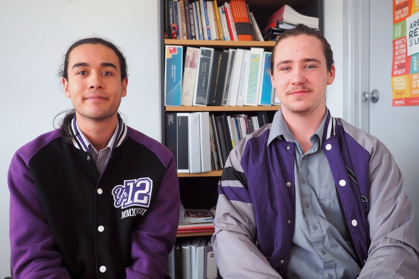 Two students in a purple and black school bomber jacket sit at a table in front of a bookshelf.