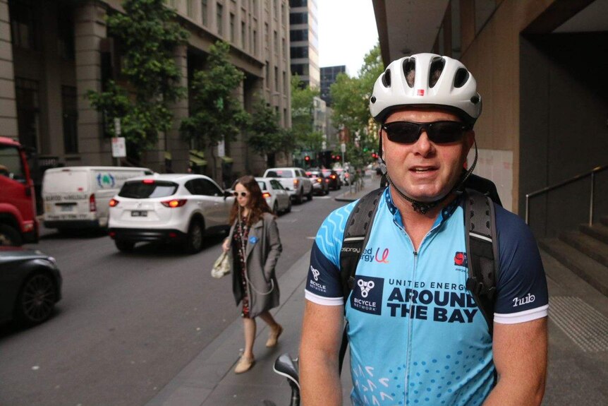 A man wearing light blue lycra and a white helmet stands with his bike on a footpath looking at the camera.