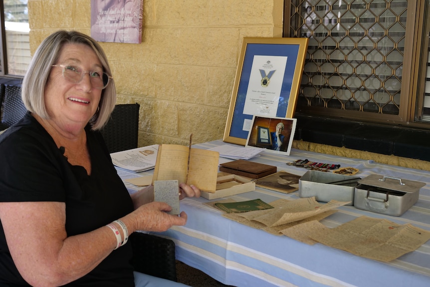 A woman sits in front of a table filled with old war memorabilia and looks at the camera smiling.