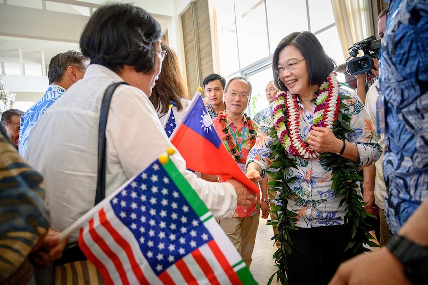 Taiwan President Tsai Ing-wen wears a necklace of flowers and shakes hands with a woman with crowd waving flags.