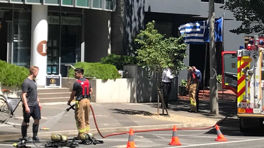 An office block with firefighter crews out front and a Greek flag waving.