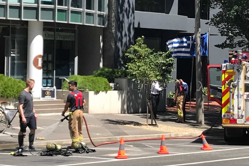 An office block with firefighter crews out front and a Greek flag waving.