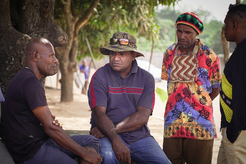 A Papua New Guinean man in a camo-print bucket hat looks thoughtful while sitting with two men under a treet