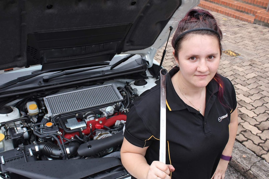 A woman in a black polo shirt holds a spanner and stands in front of a car with the bonnet open
