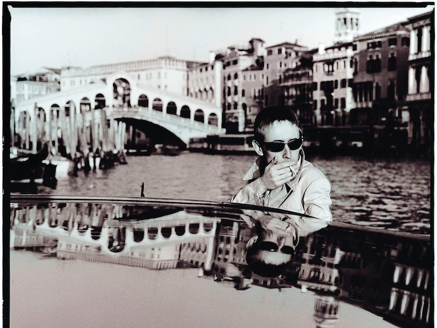 A man smokes a cigarette while riding in a boat along a river with old stone buildings and bridges behind him.