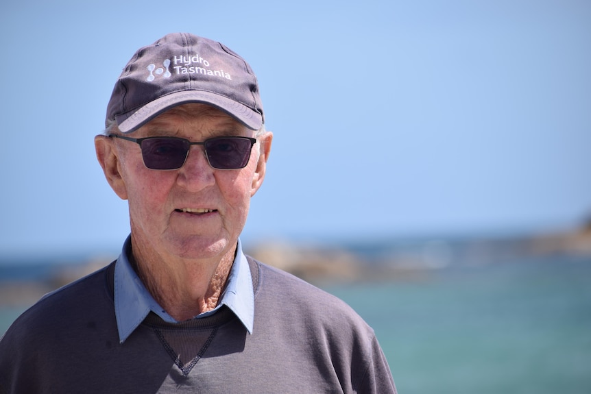 A portrait of a smiling older man wearing dark sunglasses, a blue hat and shirt. In the background is the sky and the ocean.