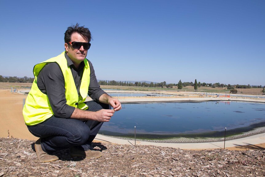 A man in a high visibility vest kneels in front of a waste water treatment pond