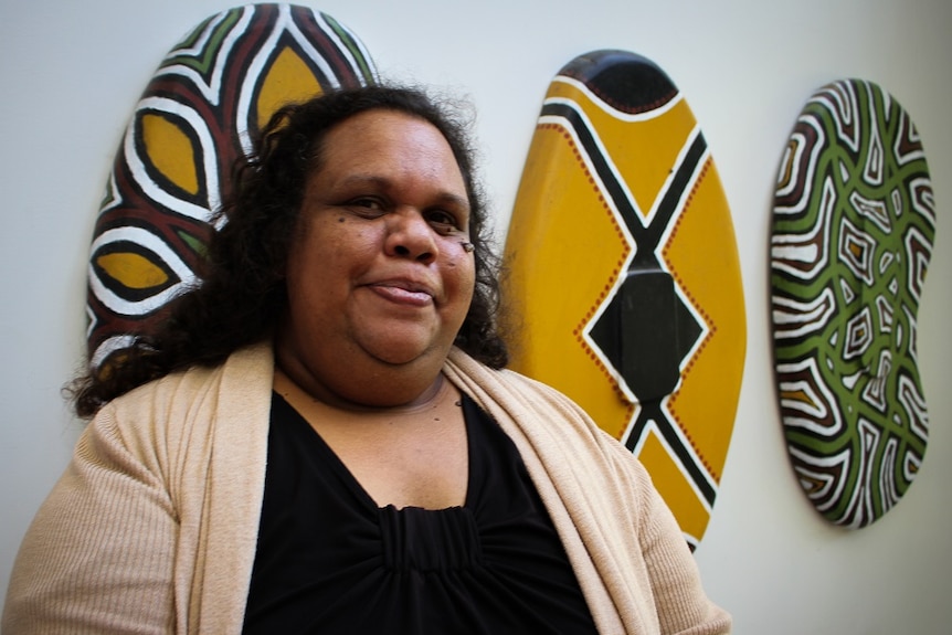 An Aboriginal woman smiles at the camera as she stands in front of three painted shields