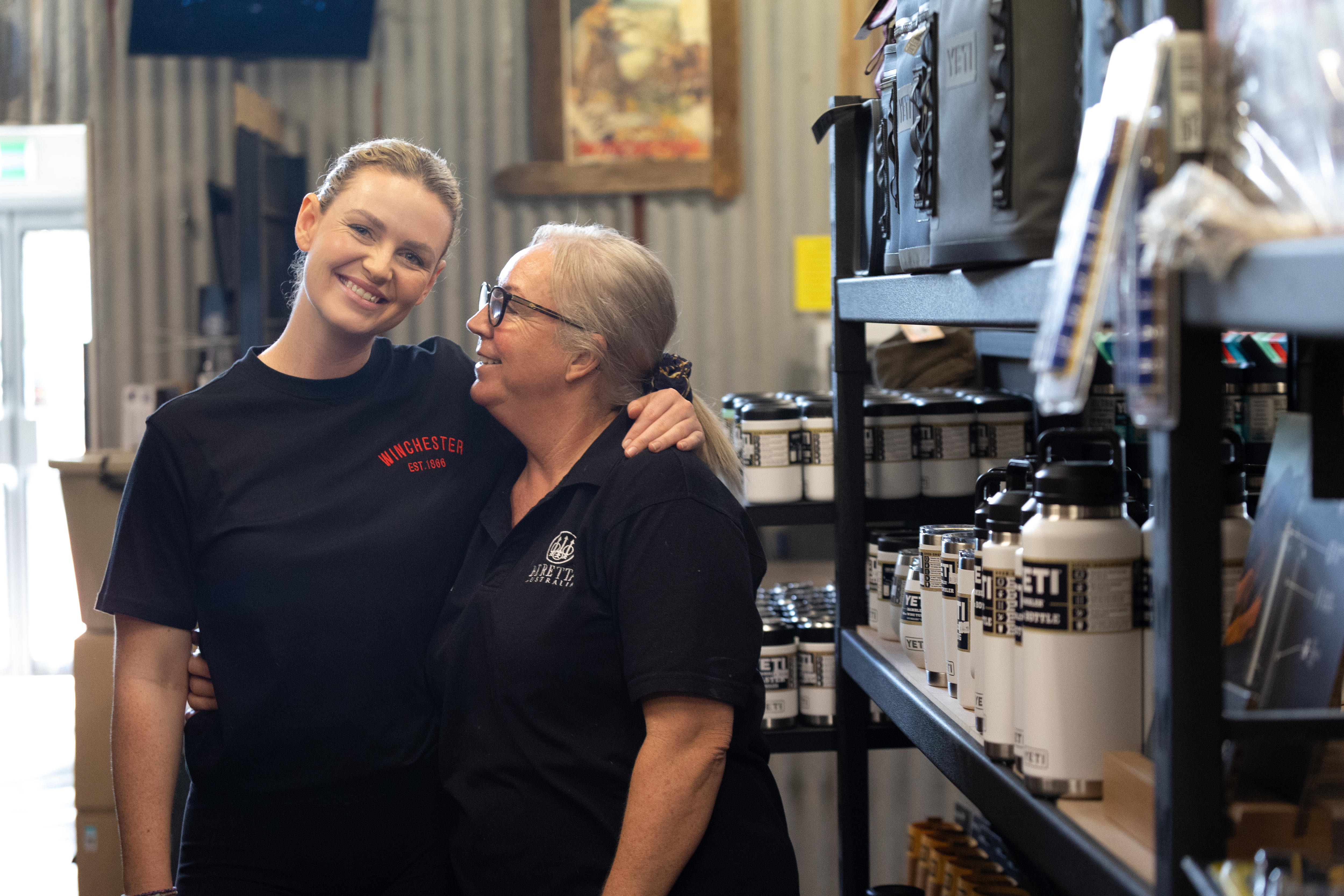 A young adult Ms Malloy smiles with one arm around her older mother who smiles at her in a storeroom with shelving.