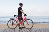 Man stands beside his red bike on a boardwalk next to the beach in Melbourne, in an article about not driving.