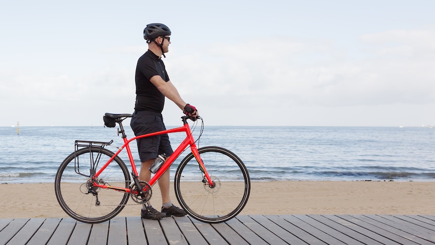 Man stands beside his red bike on a boardwalk next to the beach in Melbourne, in an article about not driving.