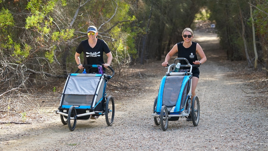 Two women push running prams through a bush track during parkrun.