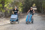 Two women push running prams through a bush track during parkrun.