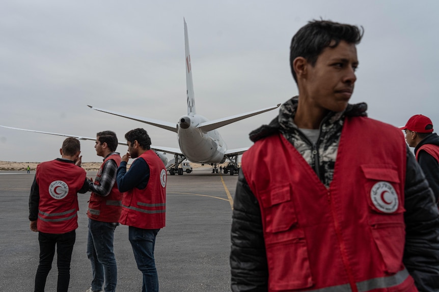 A group of aid workers wearing red jackets stand on a tarmac near a plane.