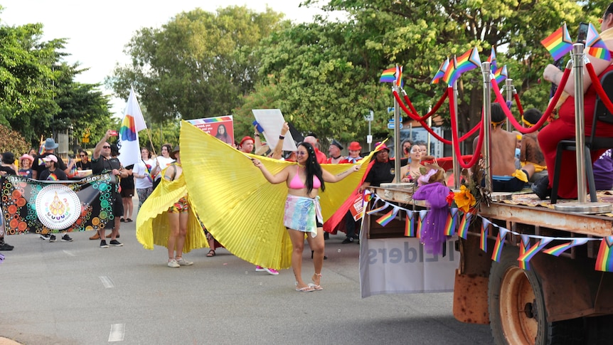 A woman walks in a parade featuring rainbow flags. She holds a large yellow drape behind her that looks like wings.