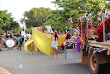 A woman walks in a parade featuring rainbow flags. She holds a large yellow drape behind her that looks like wings.