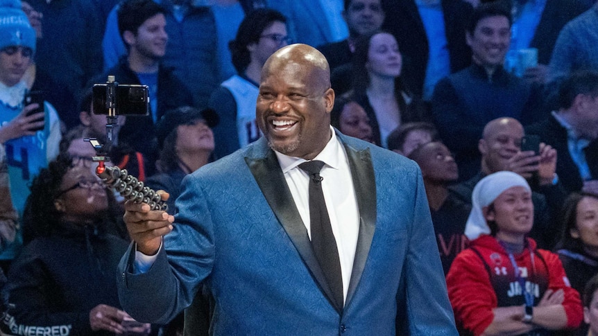 A man smiles on stage in a suit staring at a self stick with a mobile camera.