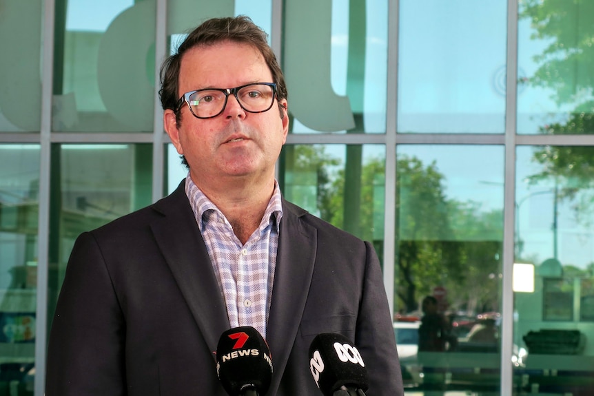 A bespectacled man with dark hair, wearing a dark suit, stands in front of a glass facade and speaks to the media.