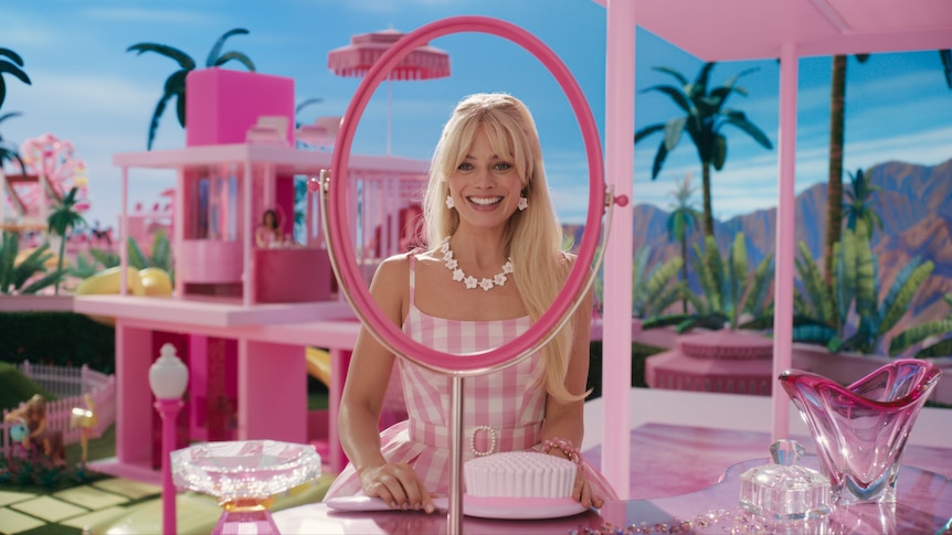 The actor Margot Robbie in a pink dress and long blonde hair playing the character Barbie in the movie, standing in a pink set