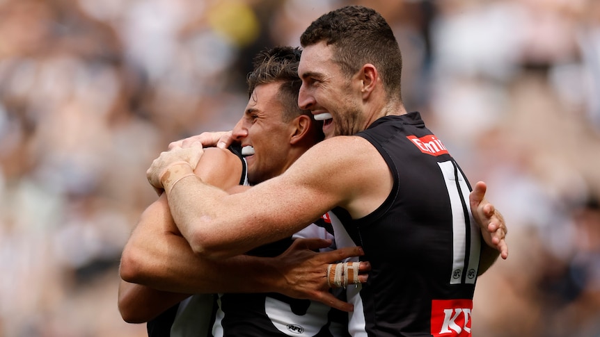 Three Collingwood AFL players embrace as they celebrate a goal.
