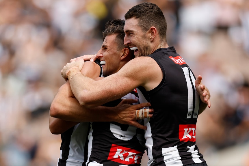 Three Collingwood AFL players embrace as they celebrate a goal.