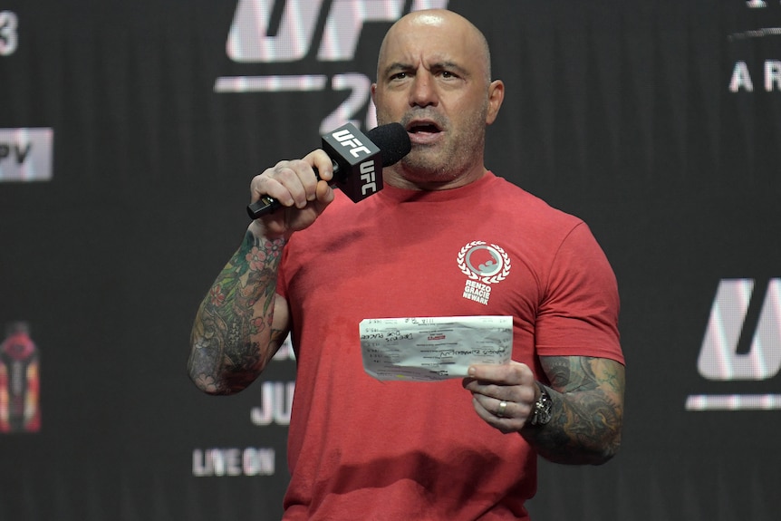 Joe Rogan holds a UFC-branded microphone up to his mouth, while reading from a piece of paper