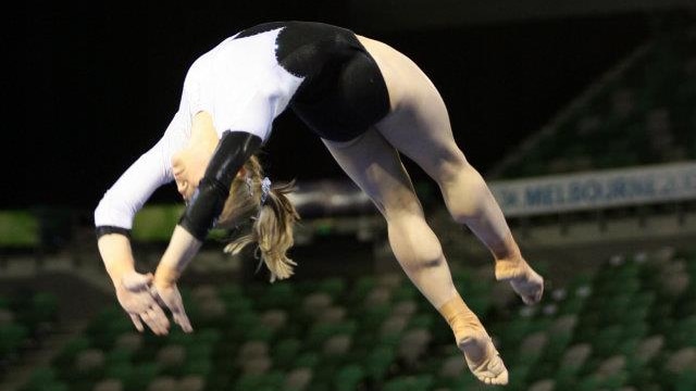 A young woman in a black and white leotard is mid-air as she flips backwards across a beam. A blurred crowd is in background.