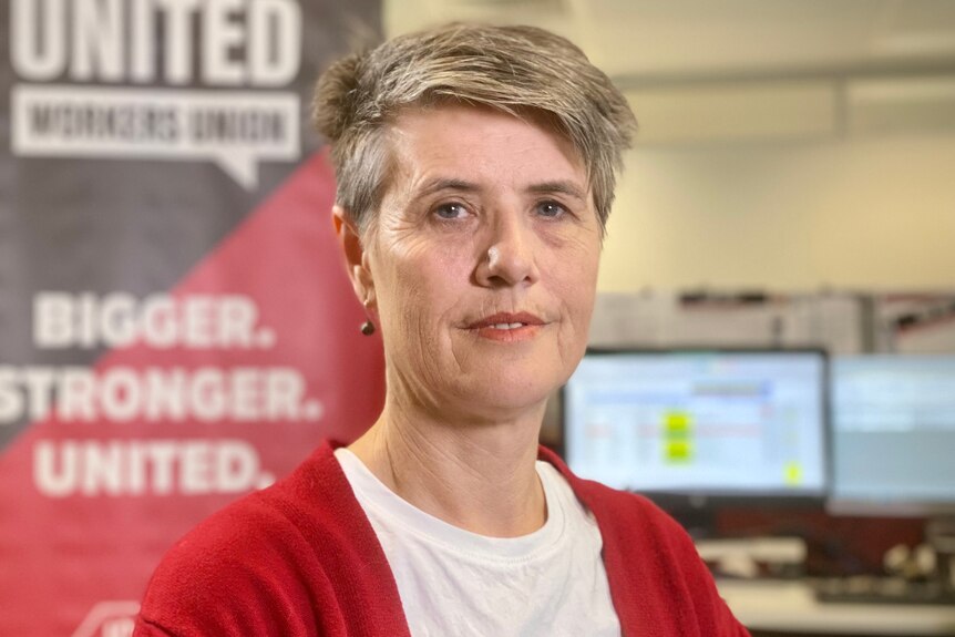 A slightly smiling short-haired woman wears a red cardigan, in front of large black and red United Workers Union sign.