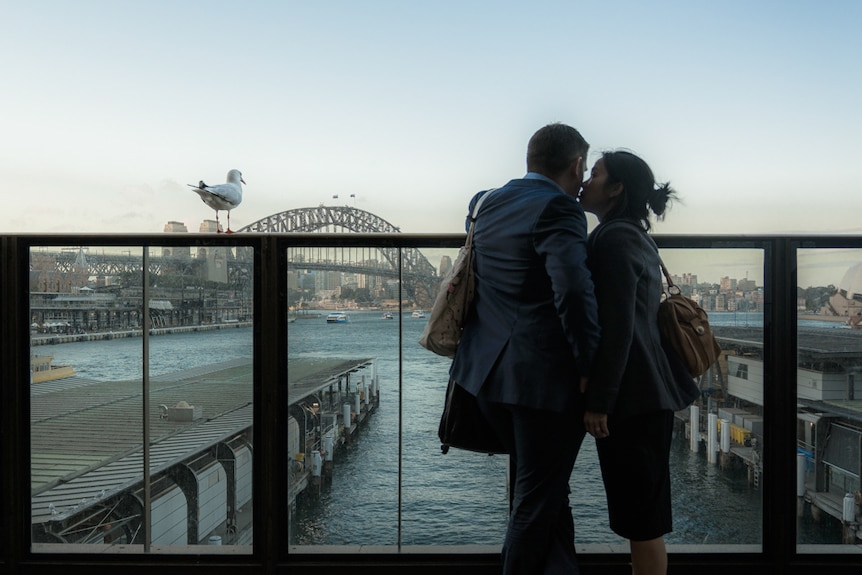 A seagull perched on a railing next to a human couple overlooking Circular Quay.