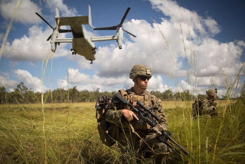 A soldier stands with a gun under a helicopter.