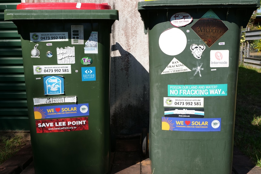 Two wheelie bins, one with red lid, the other with a yellow lid. Both are covered in save-the-environment style stickers