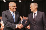 Scott Morrison, left, shakes hands with Bill Shorten, right, as they look into each other's eyes. A crowd is seated behind them.