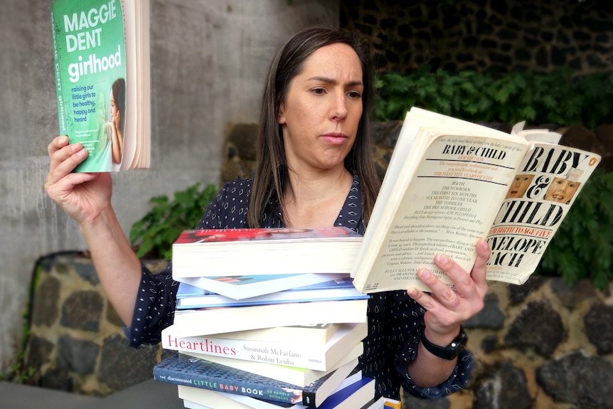A woman reads from two parenting books. A pile of more books sits in front of her.