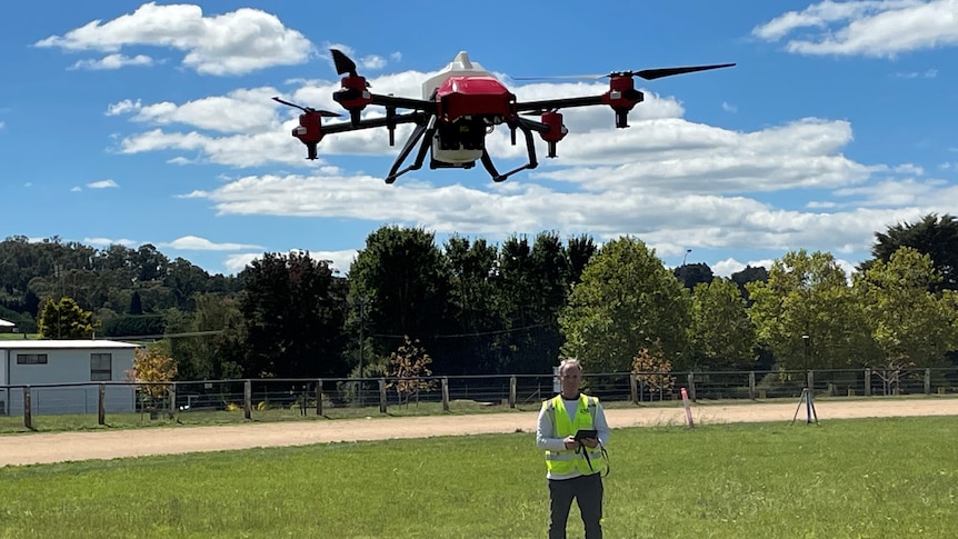 A drone in the air with a man in a yellow jacket standing in the background controlling it using a tablet.