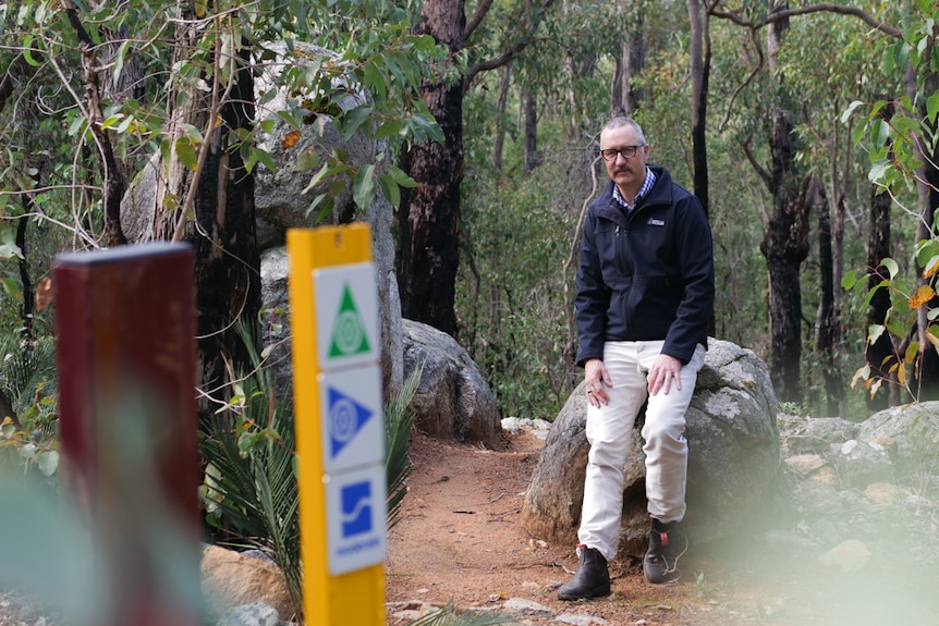 A man sitting on a rock in a forest with a mountain bike trail sign post blurred in the foreground